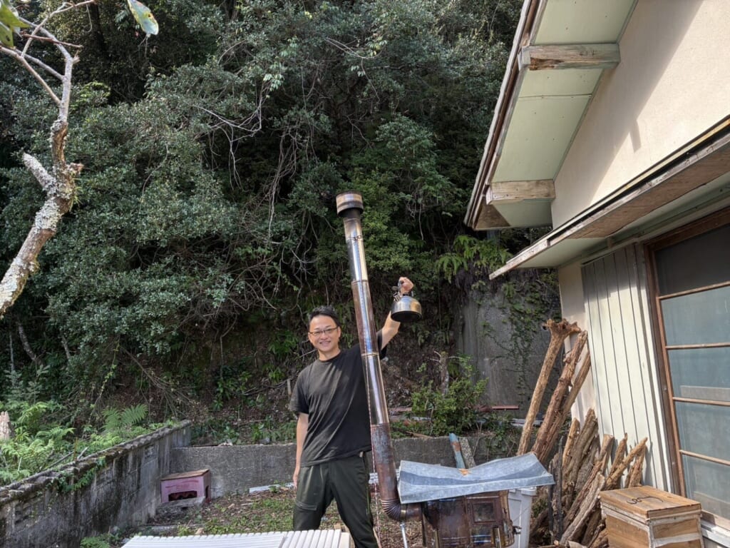 家の庭で薪ストーブの煙突の横に立ち、やかんを掲げる堀口尚志さん